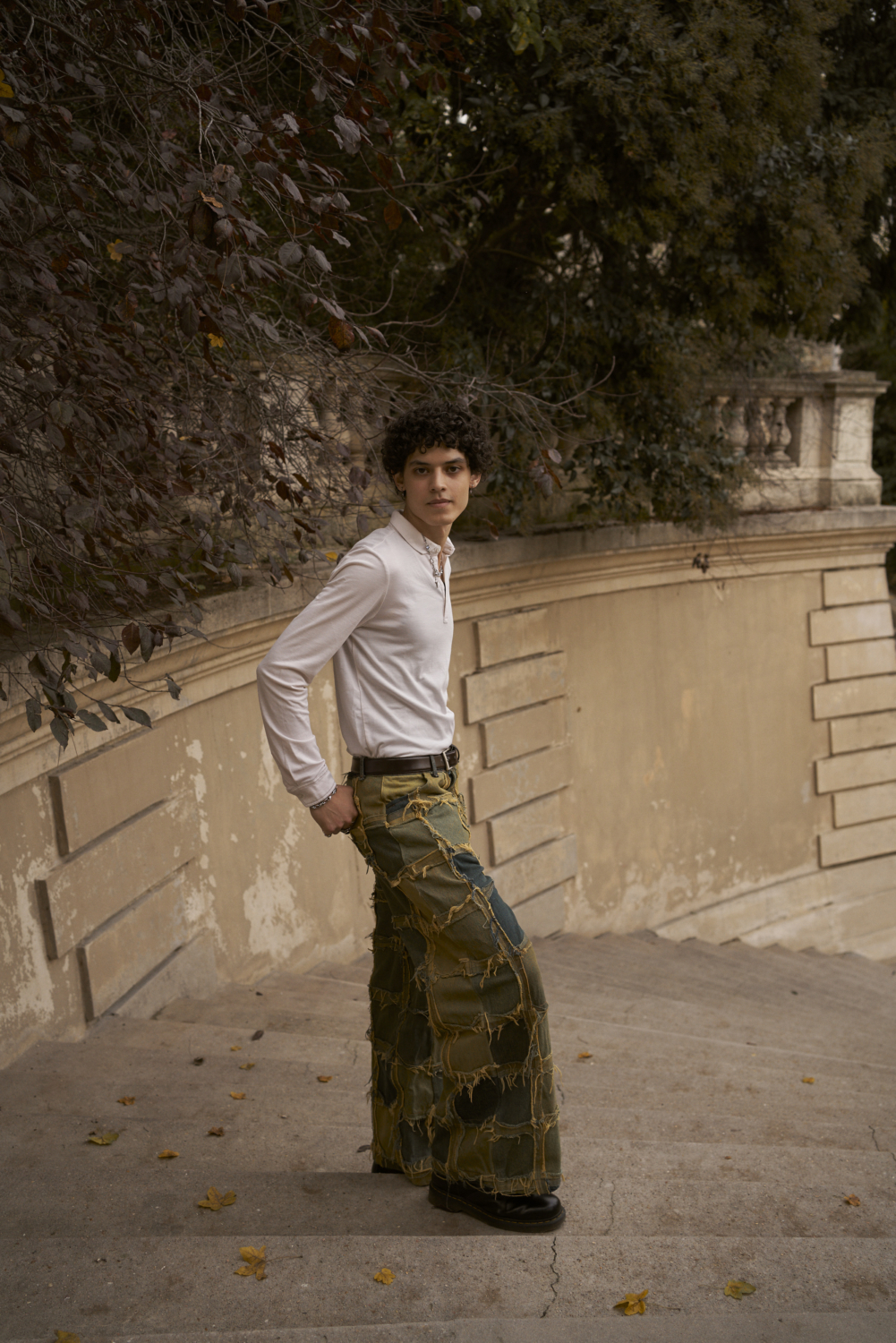 Themis in a staire case outside, wearing a white shirt, a green, yellow and blue squares patchwork pant and black doc martens